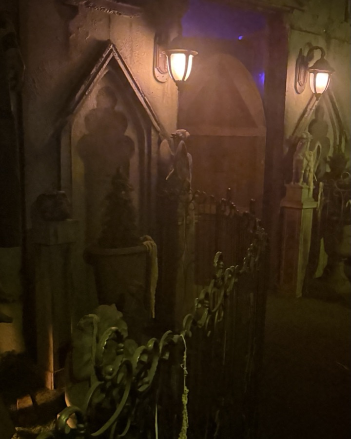 A lantern-lit stone corridor of mausoleum fronts behind an iron fence